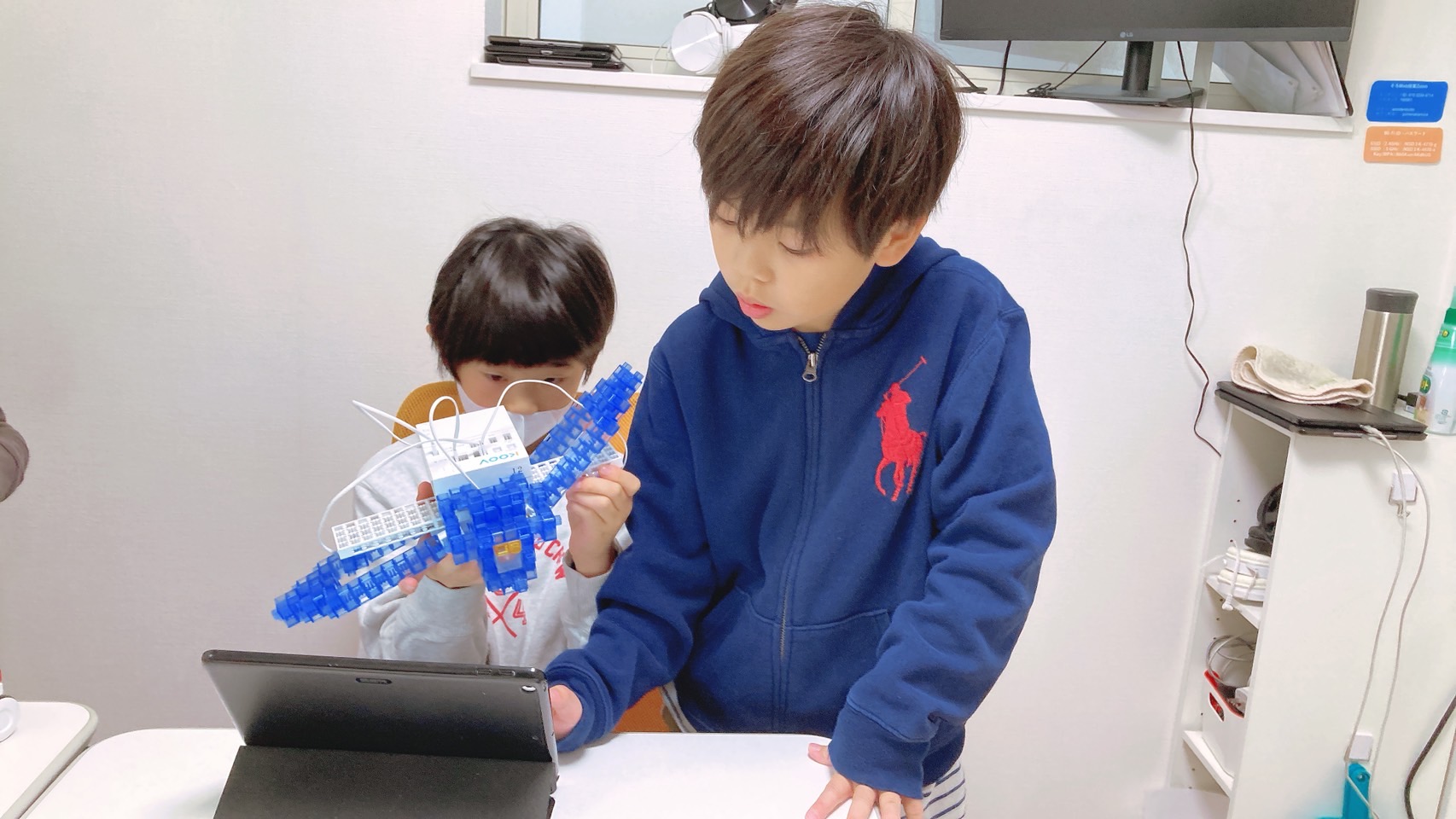 大きな青いKOOVのロボット作品と、タブレットで動きを確かめる子どもたち